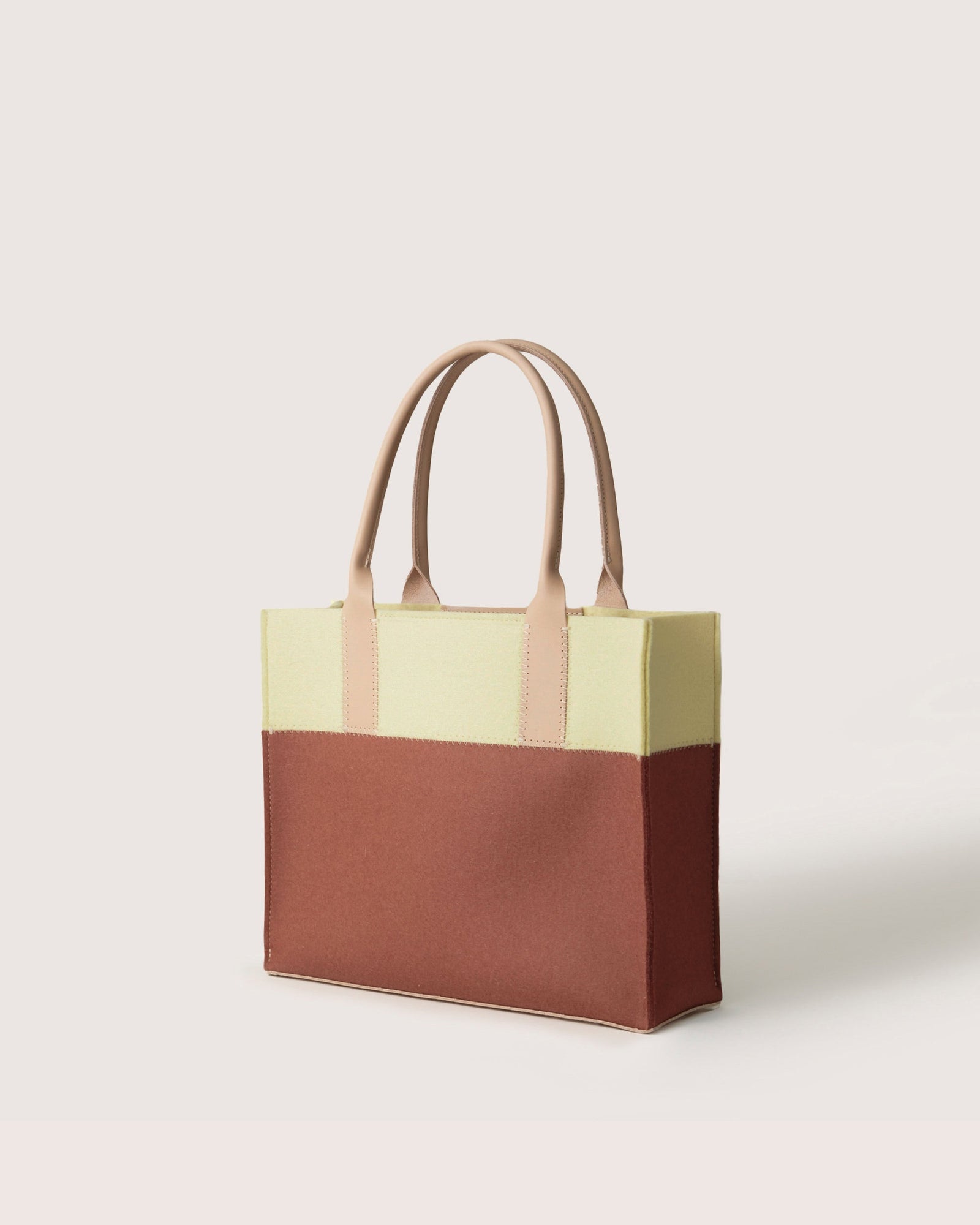 Introducing the Graf Lantz Jaunt Midi Tote, a chic mid-size carryall with a light yellow top, brown bottom, beige handles, and a white backdrop. Perfect for any occasion, this tote combines functionality and fashion effortlessly.