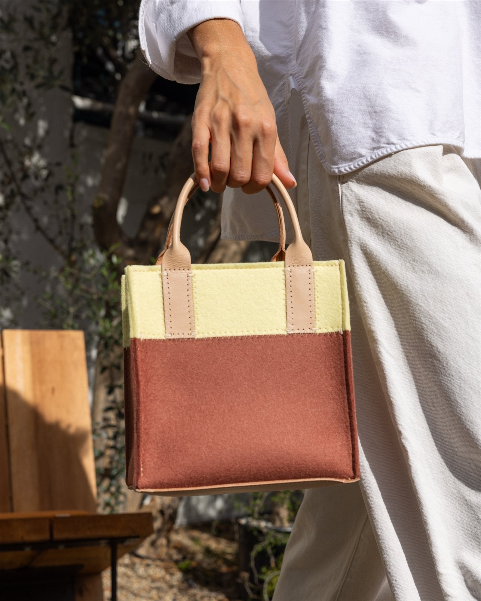 A person in a white outfit holds a Graf Lantz Mini Tote with beige handles, featuring a yellow top half and brown bottom half. A wooden chair and trees add natural charm to this stylish accessory.