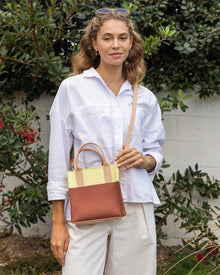 A person with wavy hair stands outdoors against a white wall and greenery, wearing a white shirt and beige pants while holding an elegant Graf Lantz mini tote. Sunglasses rest atop their head, as they gaze directly at the camera, exuding effortless style.