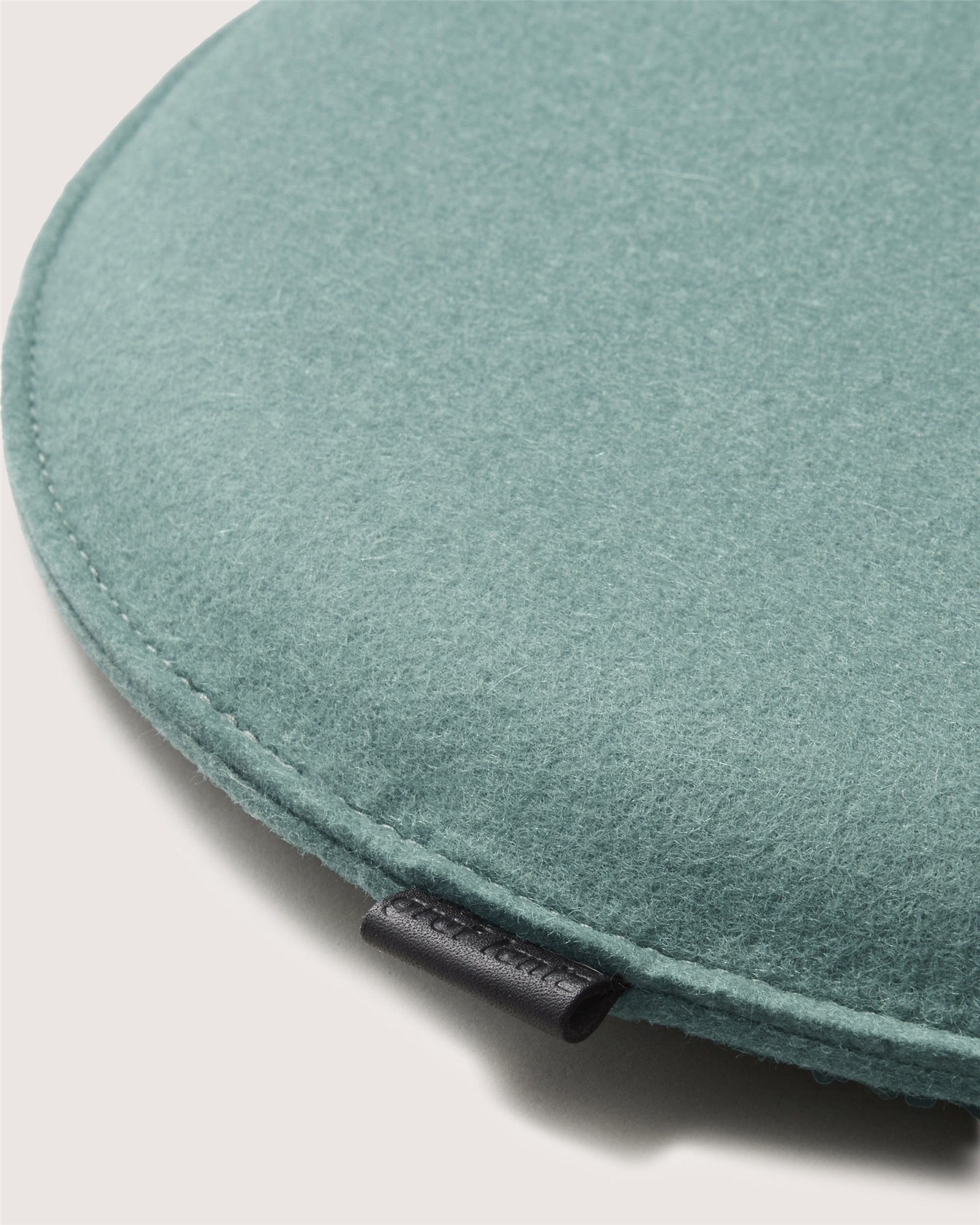 A close-up of a round, teal Graf Lantz felt meditation cushion shows soft texture, visible edge stitching, and a small black fabric tag. The light neutral background perfectly highlights this elegant Graf Lantz Zabuton Seat Cushion.