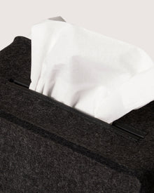 A close-up shows a white tissue partly pulled from a graf lantz dark, textured box with a minimalist silhouette akin to premium Merino wool. The plain, neutral background enhances the elegant simplicity of its design.