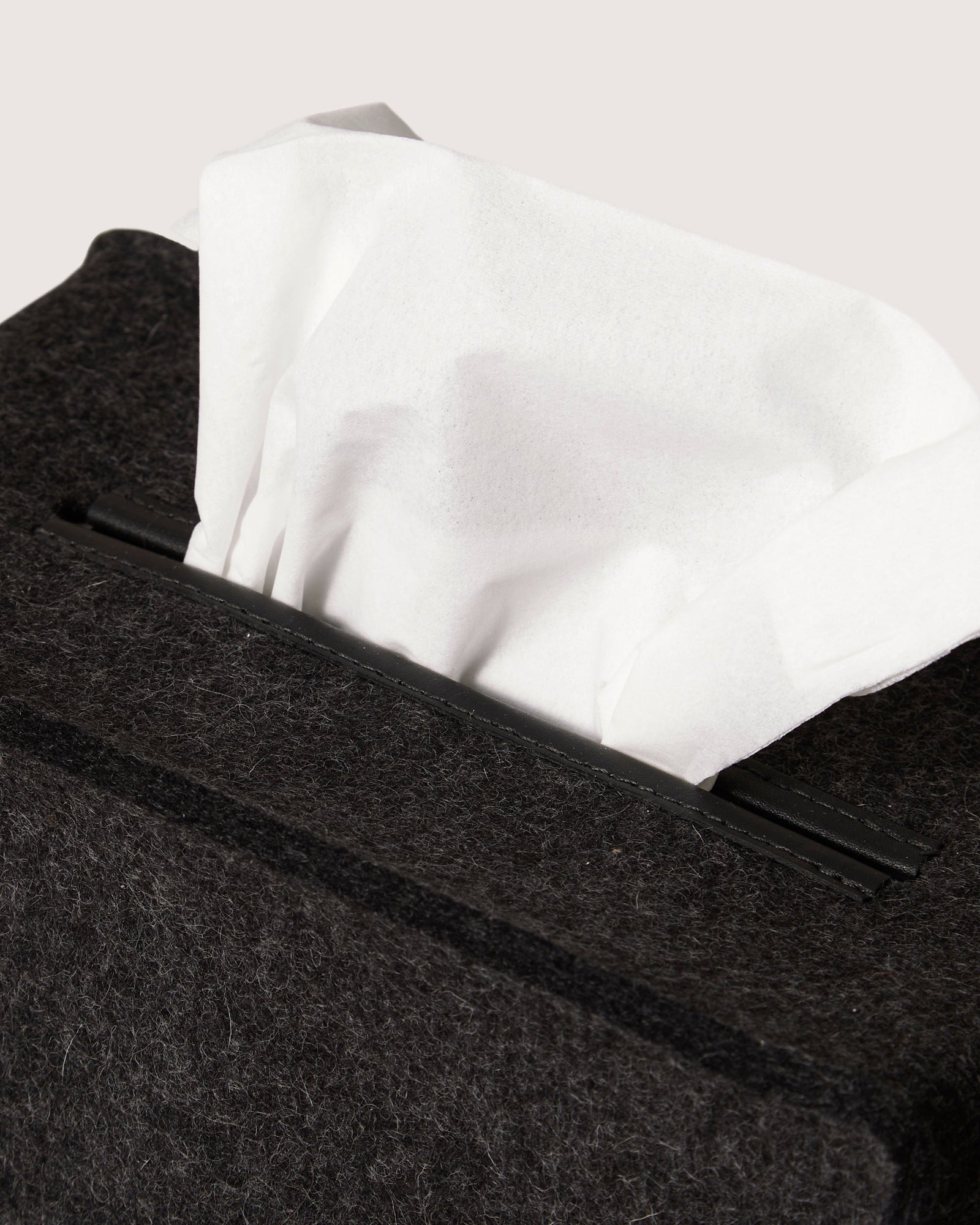 A close-up shows a white tissue partly pulled from a graf lantz dark, textured box with a minimalist silhouette akin to premium Merino wool. The plain, neutral background enhances the elegant simplicity of its design.