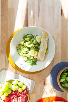 A graf lantz-crafted wooden table made from heat-resistant materials holds a salad bowl with spinach, cucumber, egg slices, olives, and bread. Nearby bowls contain green grapes, blood orange slices, and cucumber slices as sunlight casts intricate shadows.