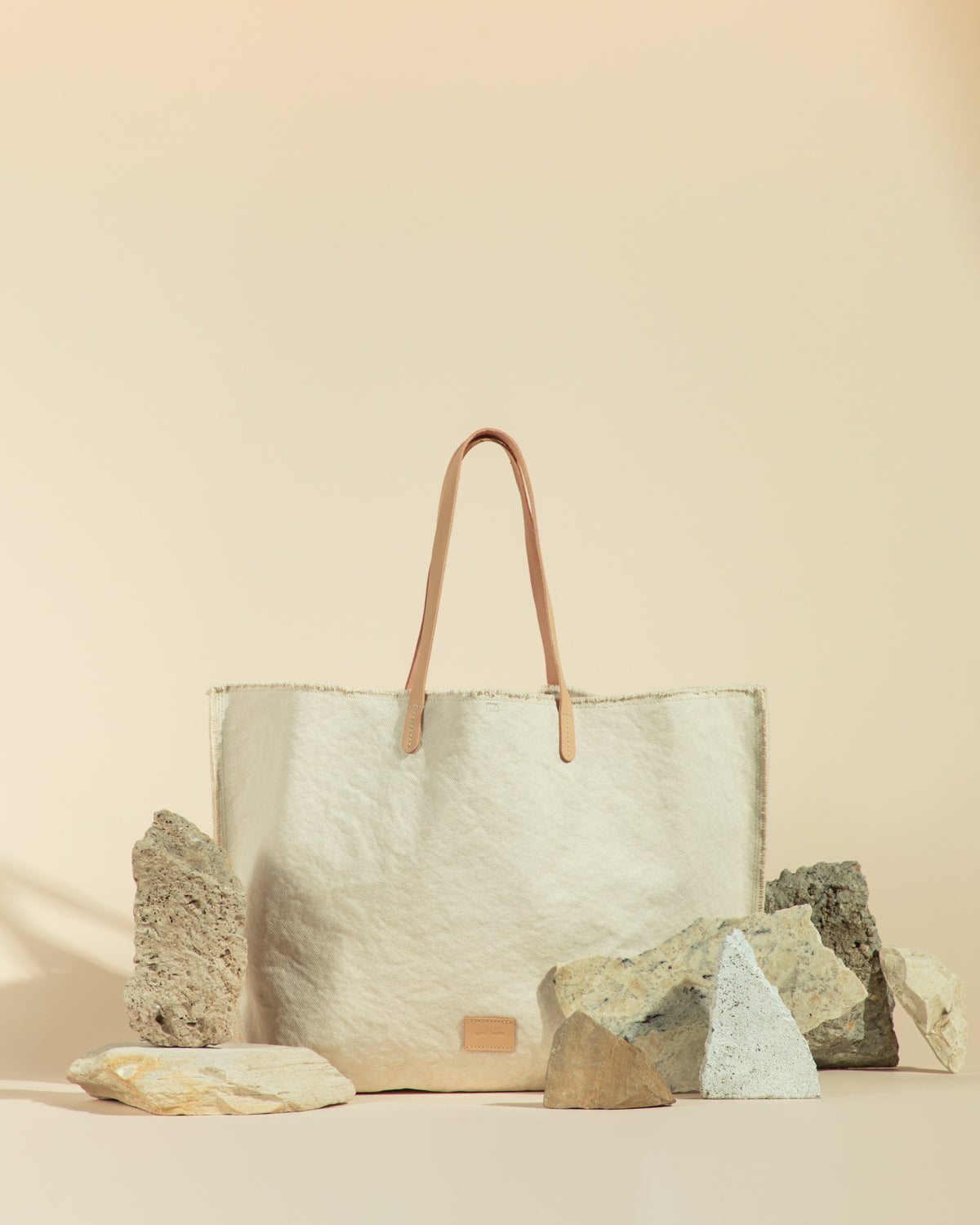 The graf lantz beige canvas tote, with its premium vegetable-tanned leather handles, is an upscale market bag. Set against a warm, light tan background with various rocks, it embodies earthy elegance and enhances its neutral tones and luxurious appeal.