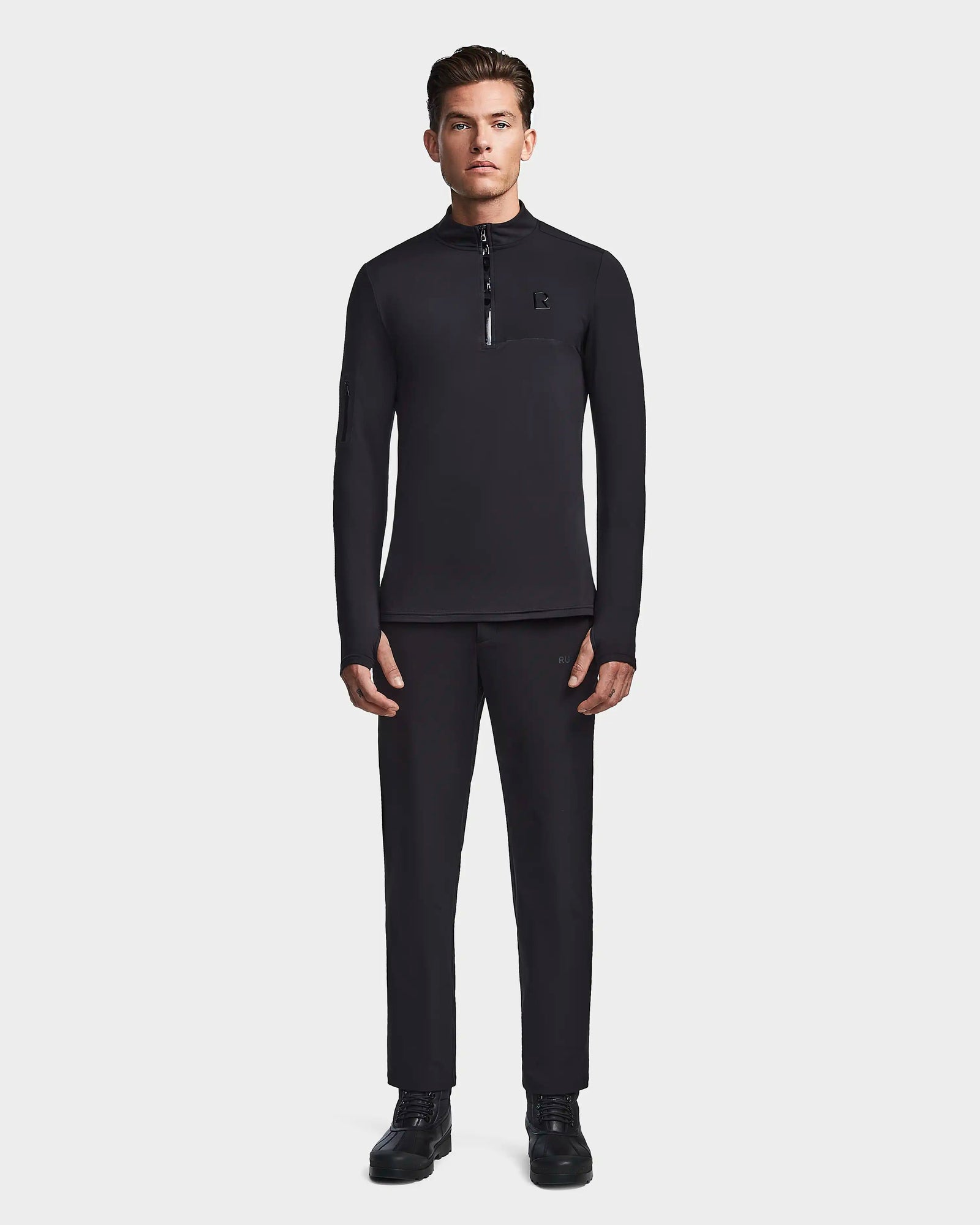 A man faces forward wearing Rudsak’s WARREN - 6523502 Black fitted stretch zip-front mock neck top and matching black pants with black shoes, set against a plain light gray background.