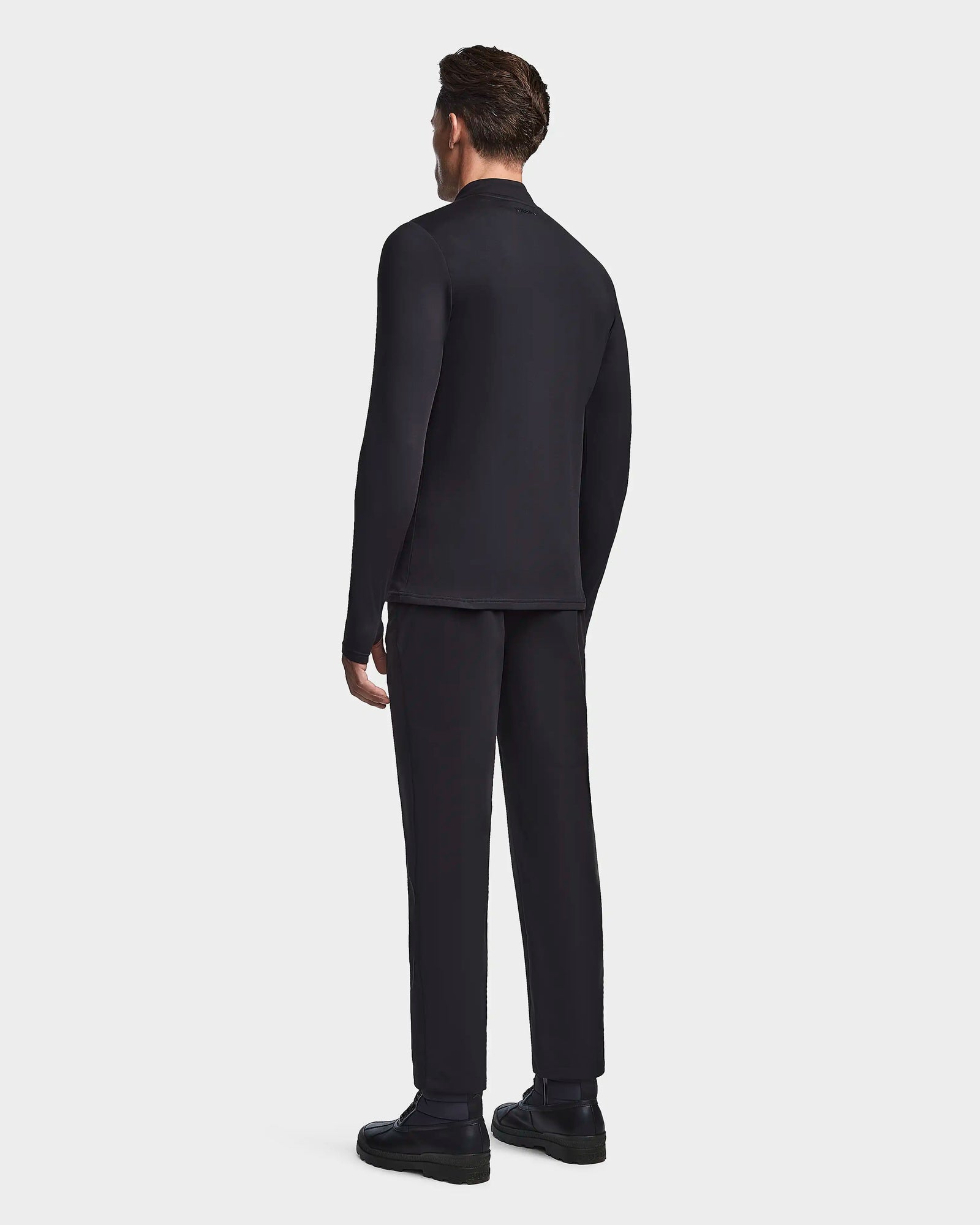 A man faces away from the camera, wearing the Rudsak WARREN - 6523502 Black mens fitted stretch top and matching pants with a zip-front mock neck, set against a plain white background.