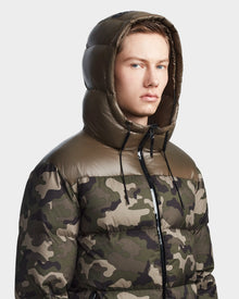 A young man models the RONNY M - M123512 Camouflage men’s hooded down puffer by Rudsak, featuring green and brown camouflage and a shiny hood, as he looks slightly to the side against a plain light background.