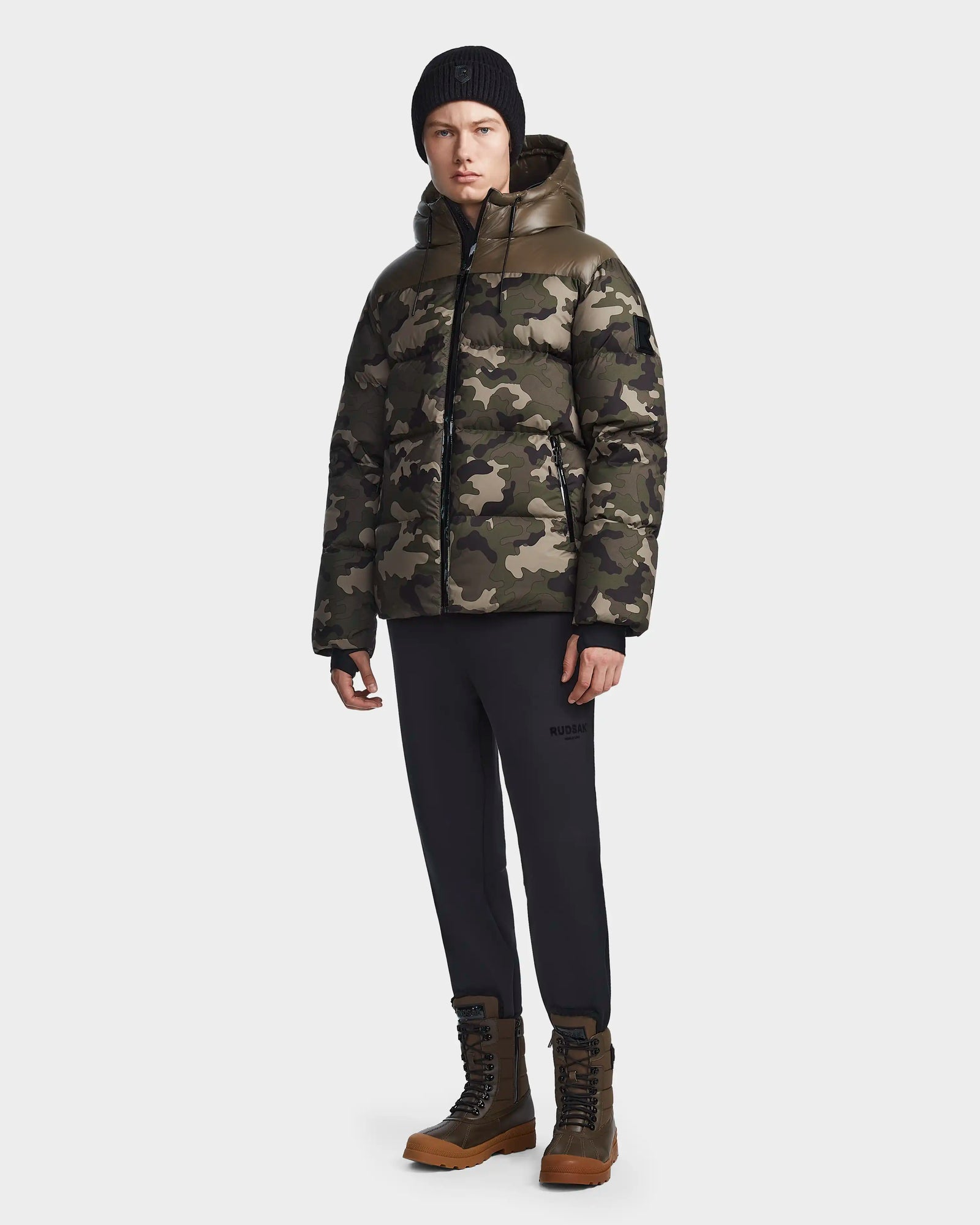 A person in the Rudsak RONNY M - M123512 Camouflage puffer jacket, black beanie, black pants, and brown lace-up boots stands facing forward against a plain light background.