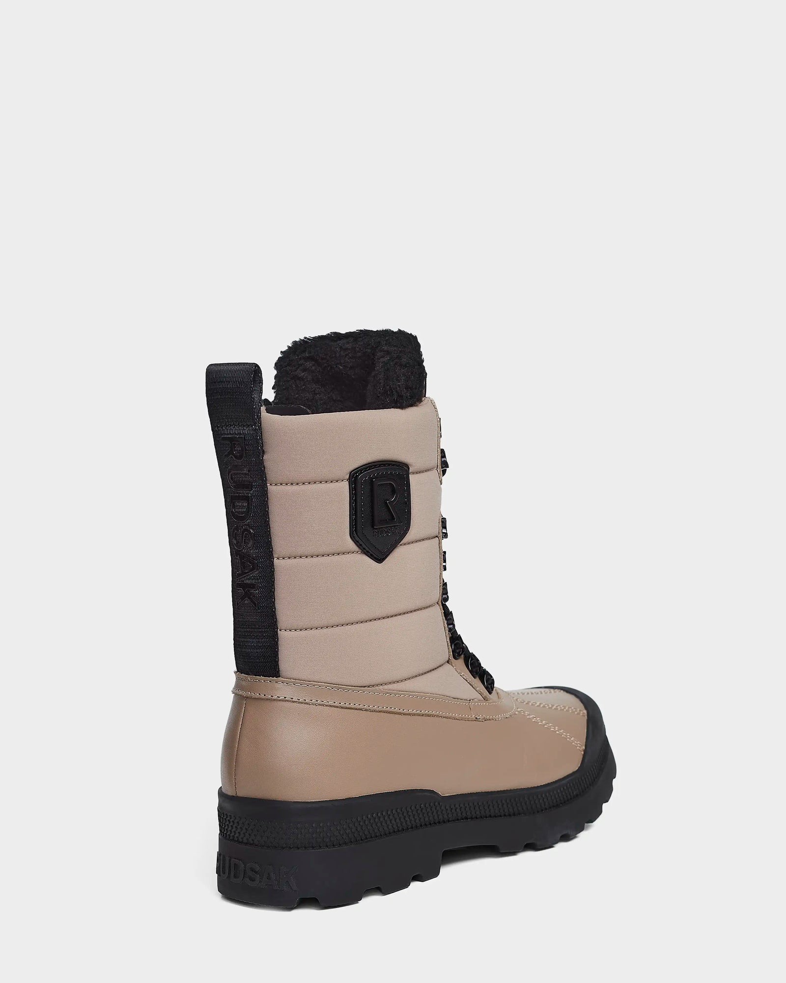 The Rudsak REAGAN - 6222500 Cement men’s winter boot features a tan and black design with thick rubber sole, black laces, padded upper, faux fur lining, and “RUDSAK” branding on the pull tab and side. Boot angled left.