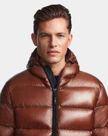 A man with short brown hair wears the Rudsak DEMIAN S - M123508 Cohiba, a shiny, water- and wind-resistant hooded down puffer jacket. It’s zipped up as he faces forward with a neutral expression against a plain light background.