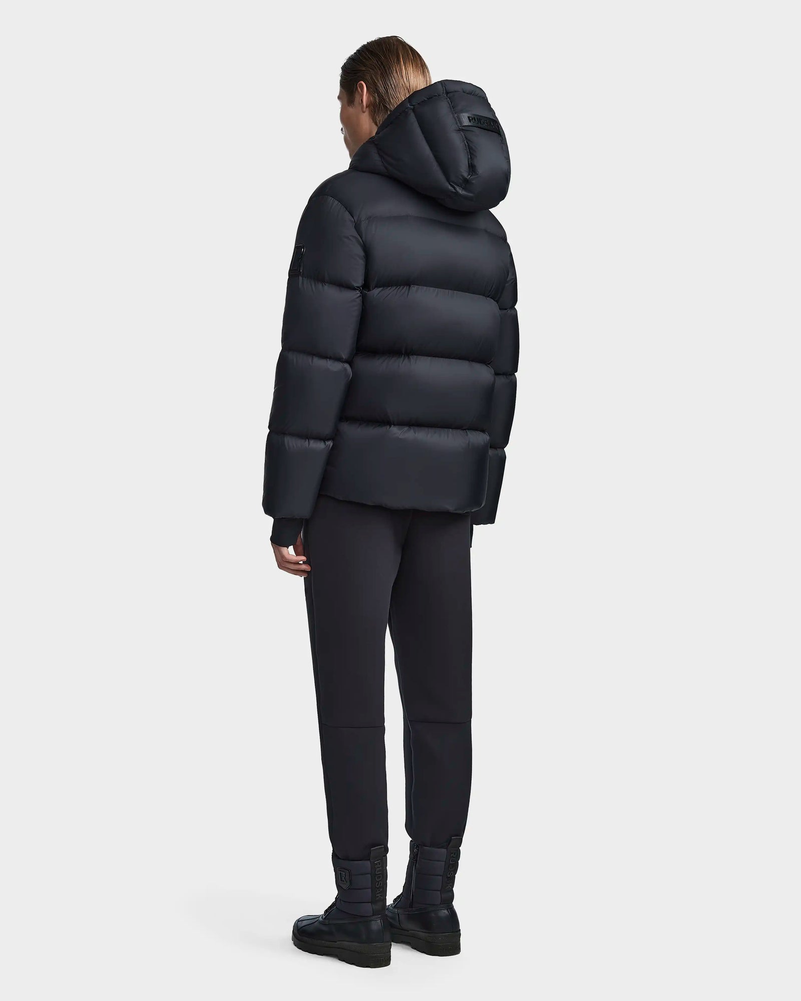A person faces away from the camera, wearing the Rudsak DANIEL M - M123510 BLACK mens hooded down puffer jacket, styled with black pants and boots against a plain light backdrop.