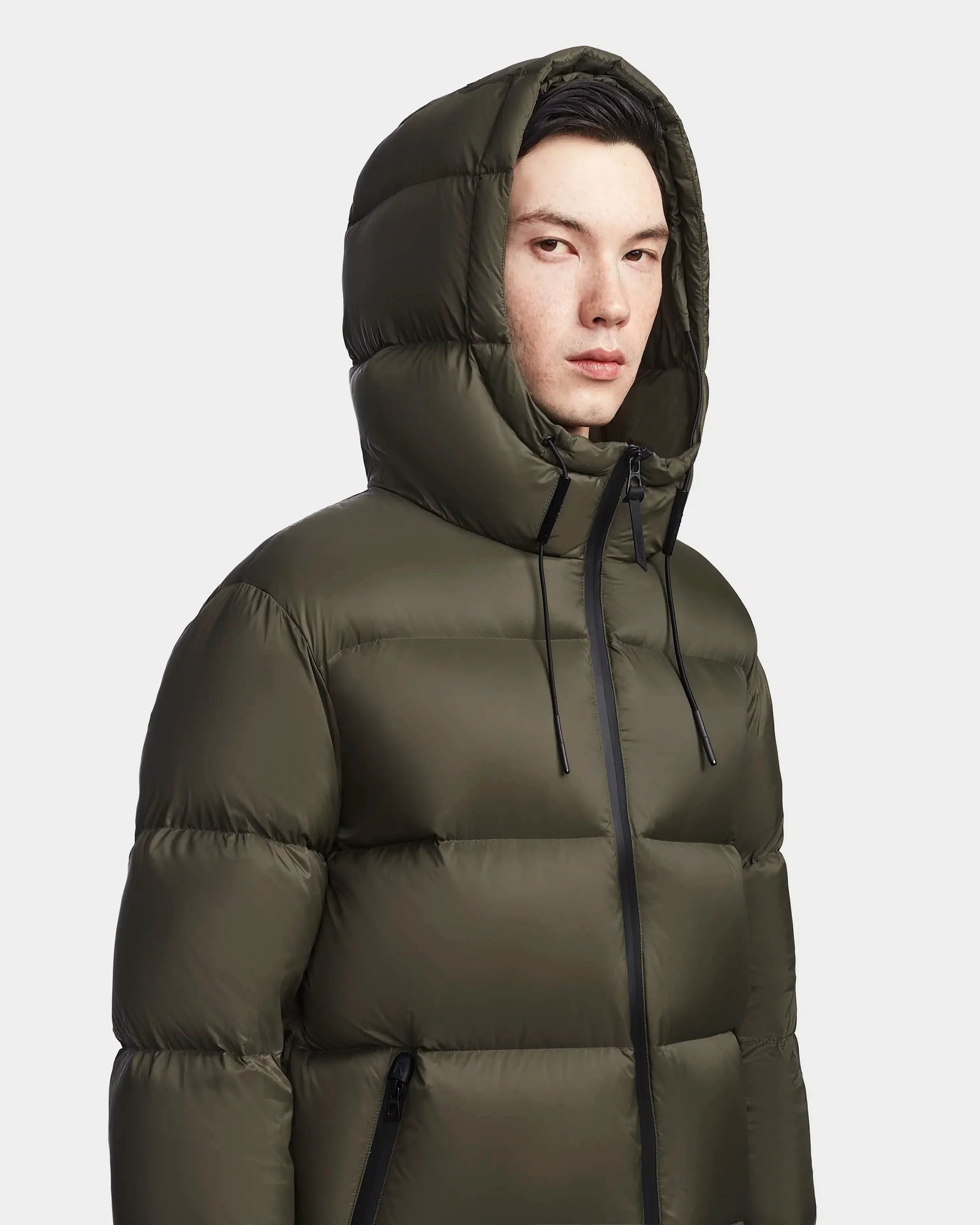 A person wearing the Rudsak DANIEL M - M123510 Light Olive weather-resistant winter jacket with a hood, standing against a plain light background and looking slightly to the side.