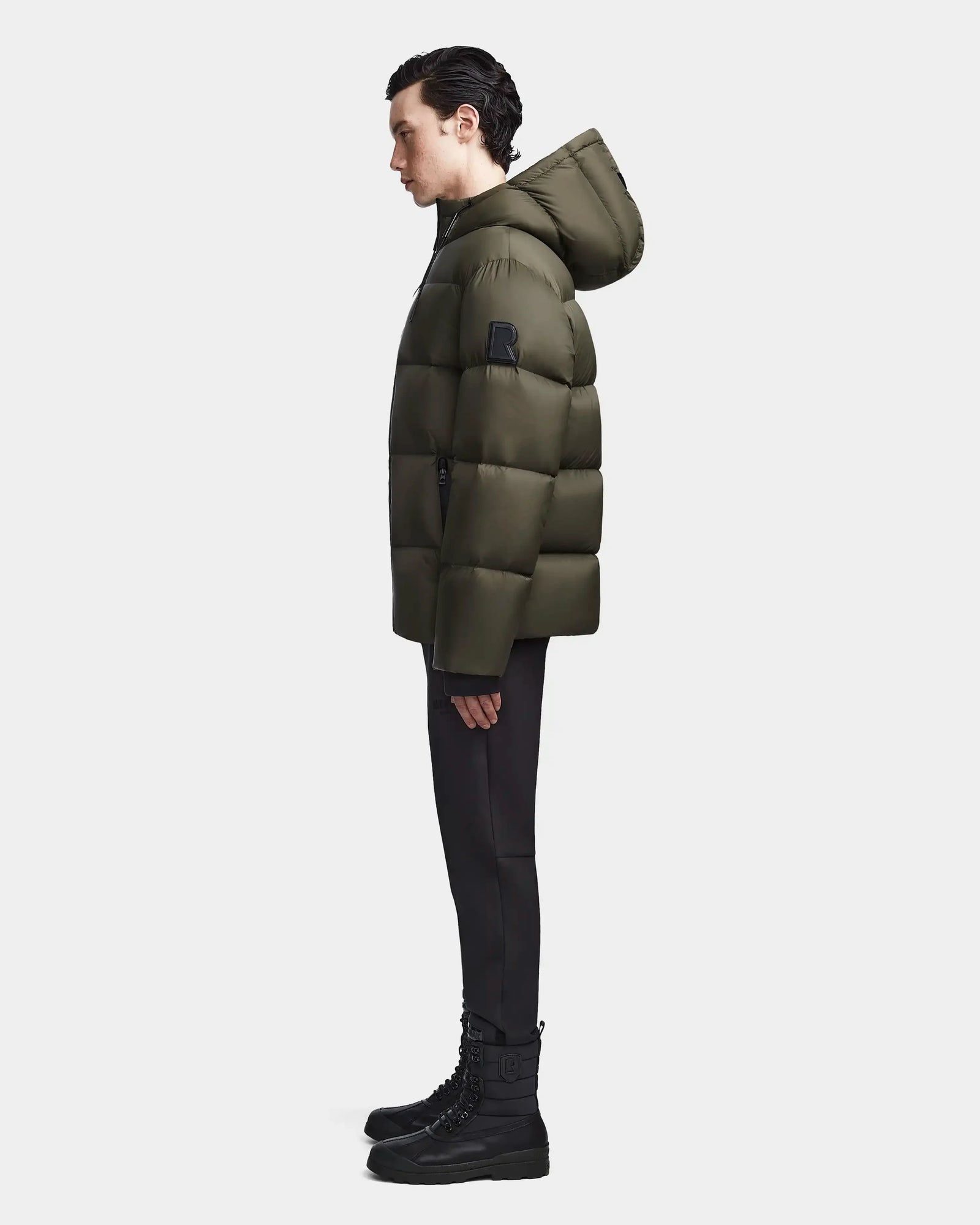 A person stands in profile against a plain background, wearing the Rudsak DANIEL M - M123510 Light Olive mens coat with a hood, paired with black pants and black lace-up boots.