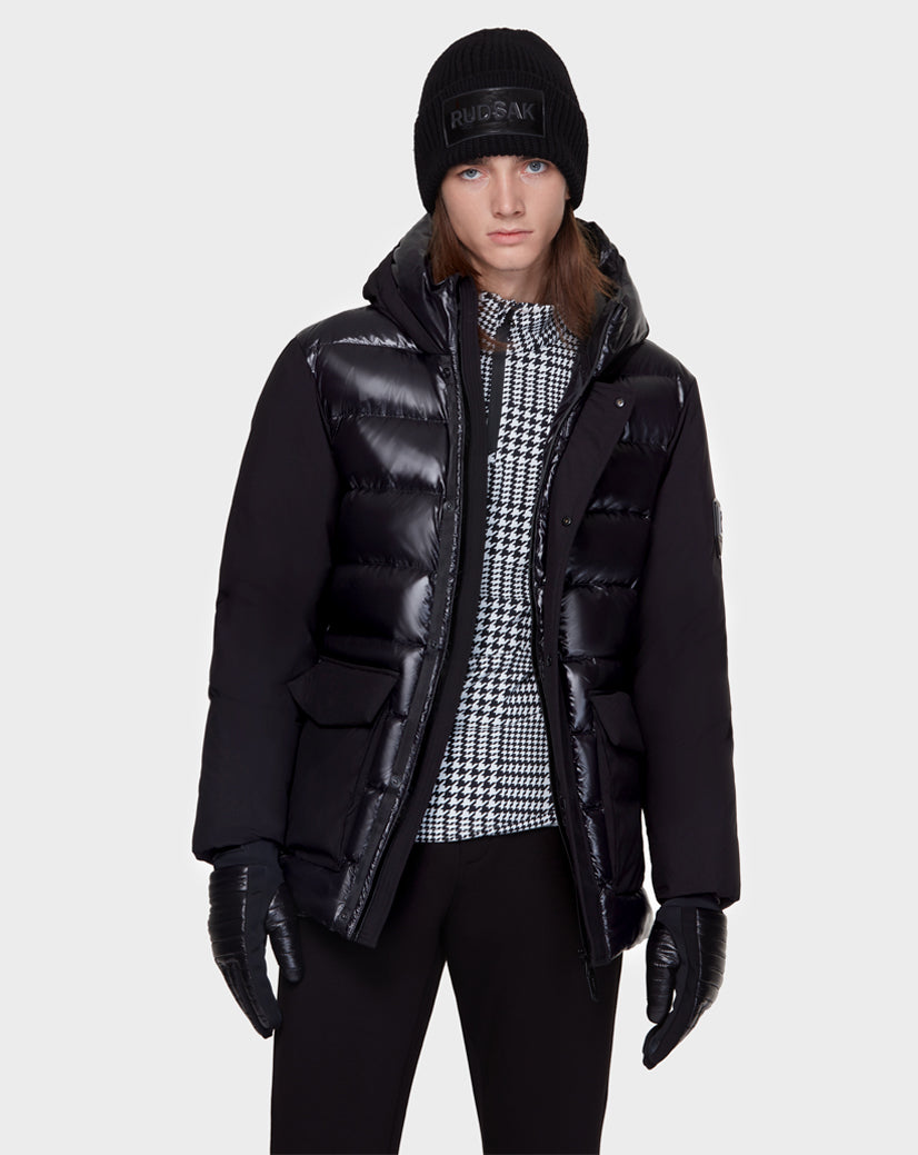 A person with straight brown hair wears the Rudsak DUSK - M121506 BLACK men’s puffer jacket, a houndstooth-patterned shirt, black beanie, gloves, and pants, standing against a plain light gray background.