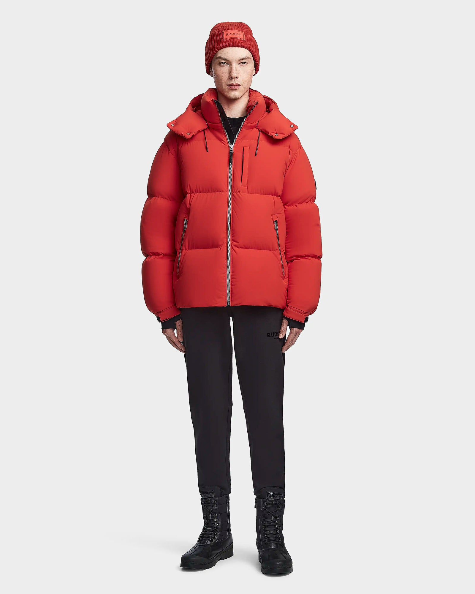 A person wearing the VINCENT M - 6123510 Orange down puffer jacket by Rudsak, a red beanie, black pants, and black lace-up boots stands facing forward against a plain white background.