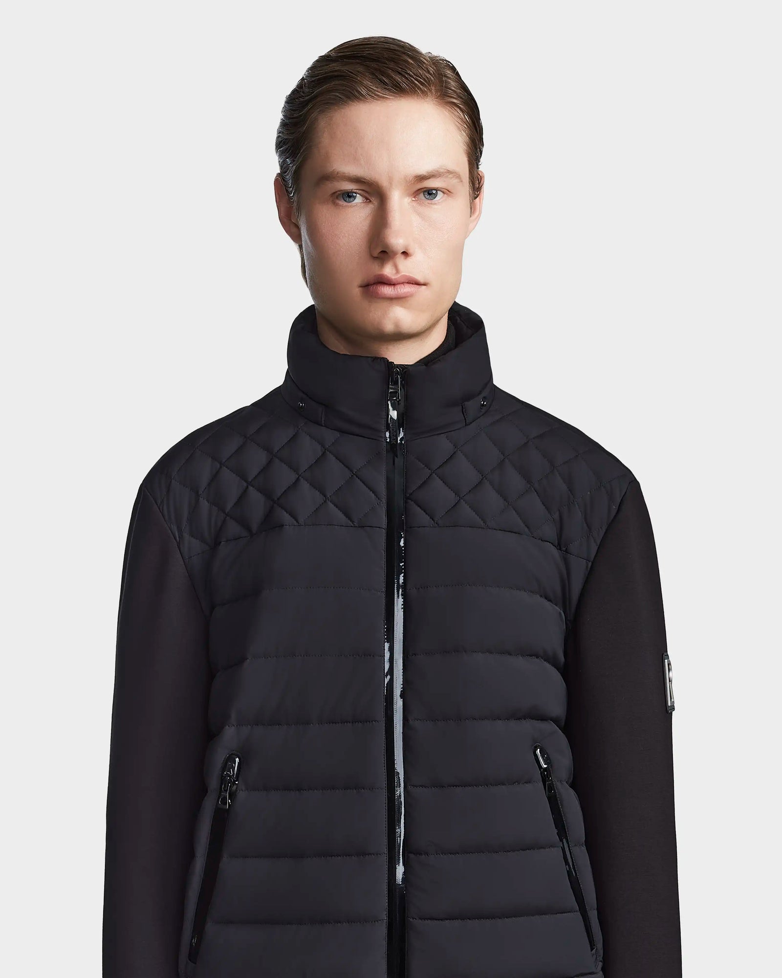 A young man with fair skin and straight light brown hair wears the Rudsak LAURENZO - M123514 Black men’s light down jacket with a high collar, standing facing forward against a plain light gray background.