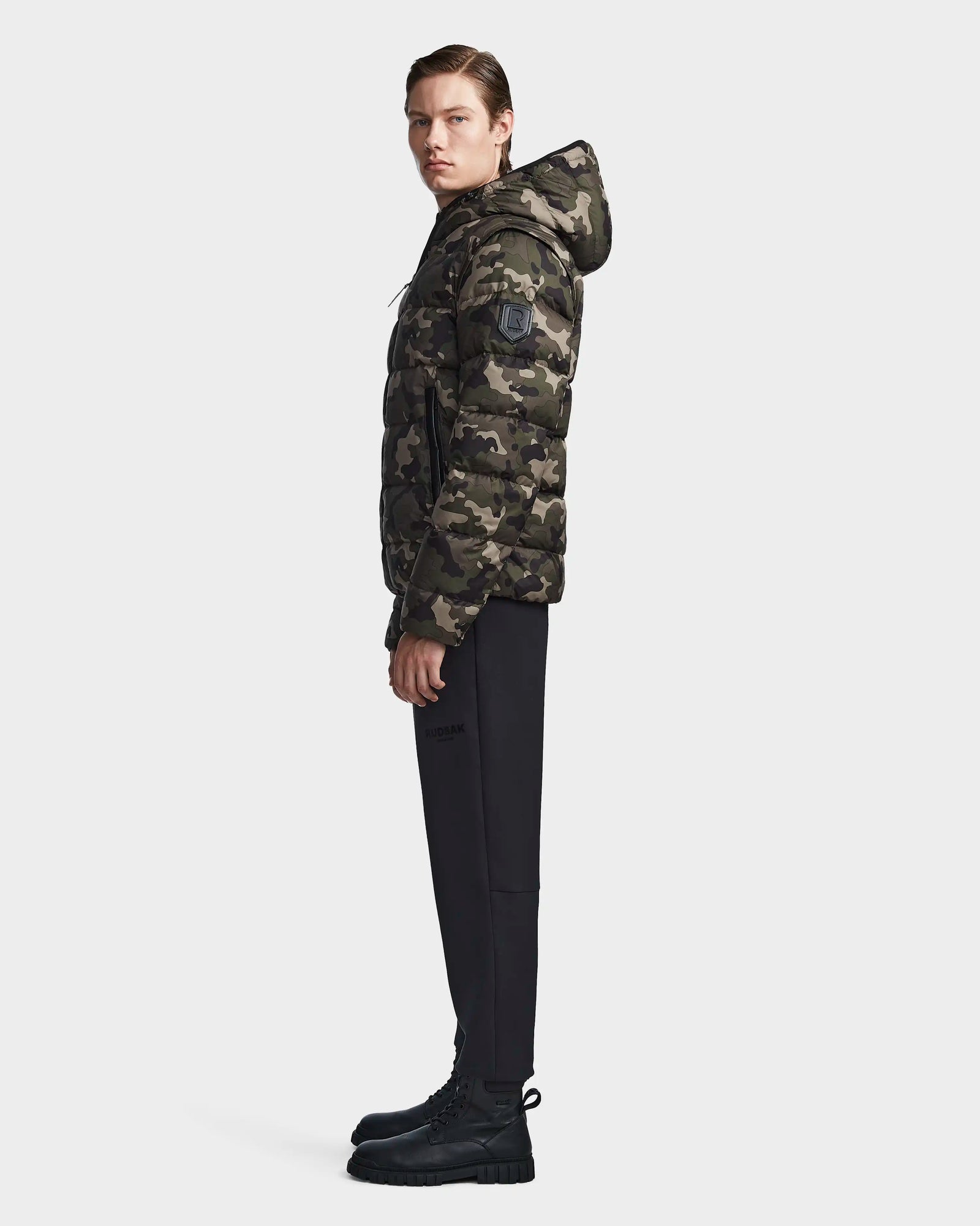 A person stands in profile wearing the Rudsak DEMIAN M-R - M123504 Camouflage mens down puffer hooded jacket, paired with black pants and black boots, against a plain white background.