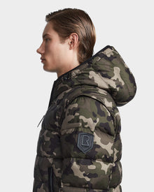 A young man with straight, light brown hair models Rudsaks DEMIAN M-R - M123504 Camouflage down puffer in green and brown, made from recycled fabric, with a black R patch on the sleeve, shown in profile against a white background.