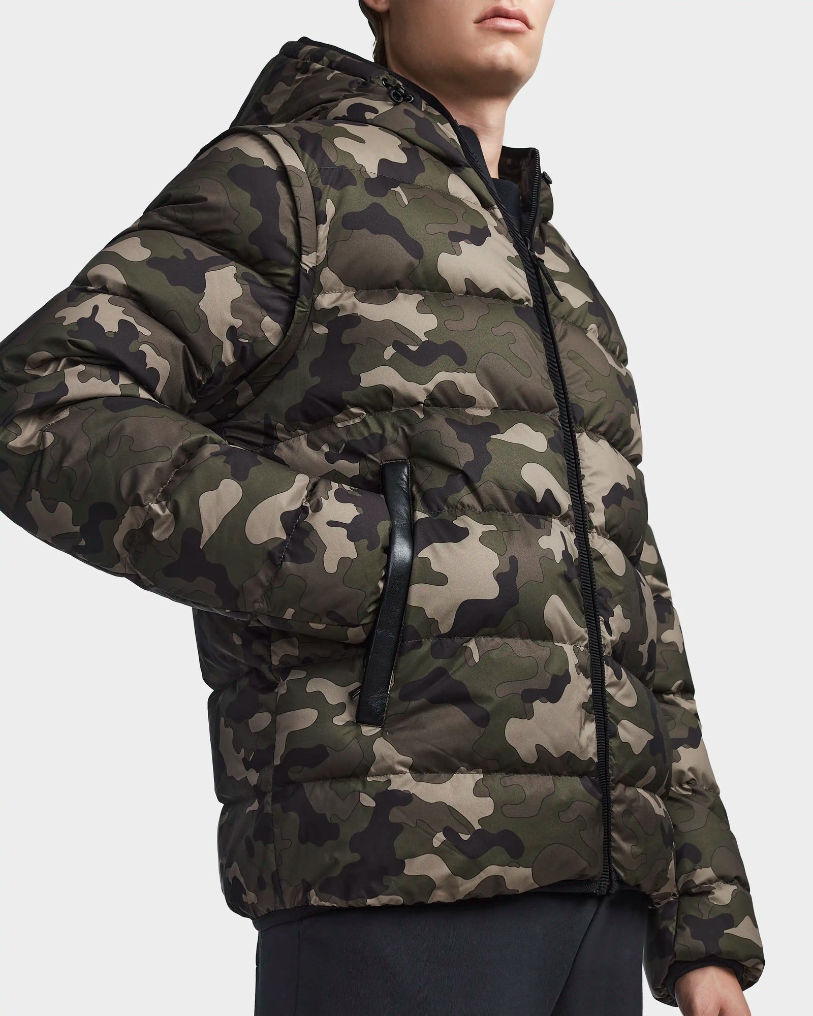 A person wears the Rudsak DEMIAN M-R - M123504 Camouflage men’s down puffer jacket, featuring a green, brown, and black camo print, hood, black zipper, and side pockets. The face is partially out of frame. Made from sustainable recycled fabric.
