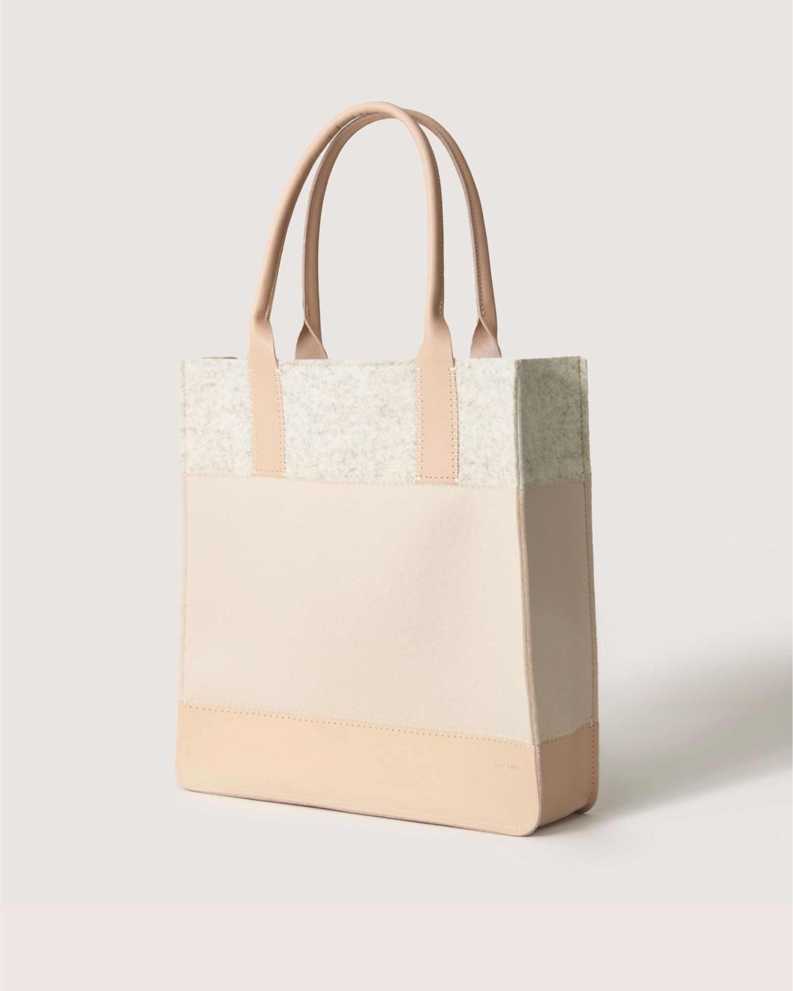 Introducing the Graf Lantz Jaunt Midi Tote, a chic mid-size beige and cream carryall. It features two sturdy handles, a minimalist design, textured upper, smooth bottom, and is crafted for detail. This elegant bag stands upright on any surface.