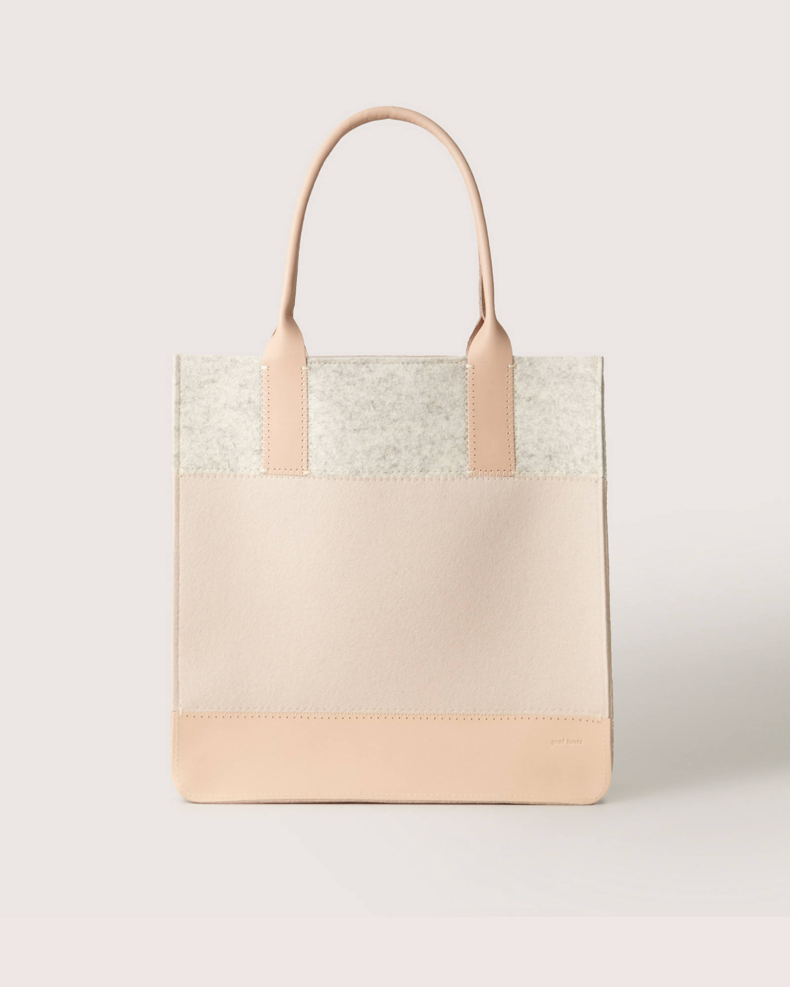 The Graf Lantz Jaunt Midi Tote is a chic, mid-size carryall in light beige and cream blocks. It features two sturdy handles and is crafted from luxurious Merino wool, offering a simple, modern design that stands out beautifully against a plain background.