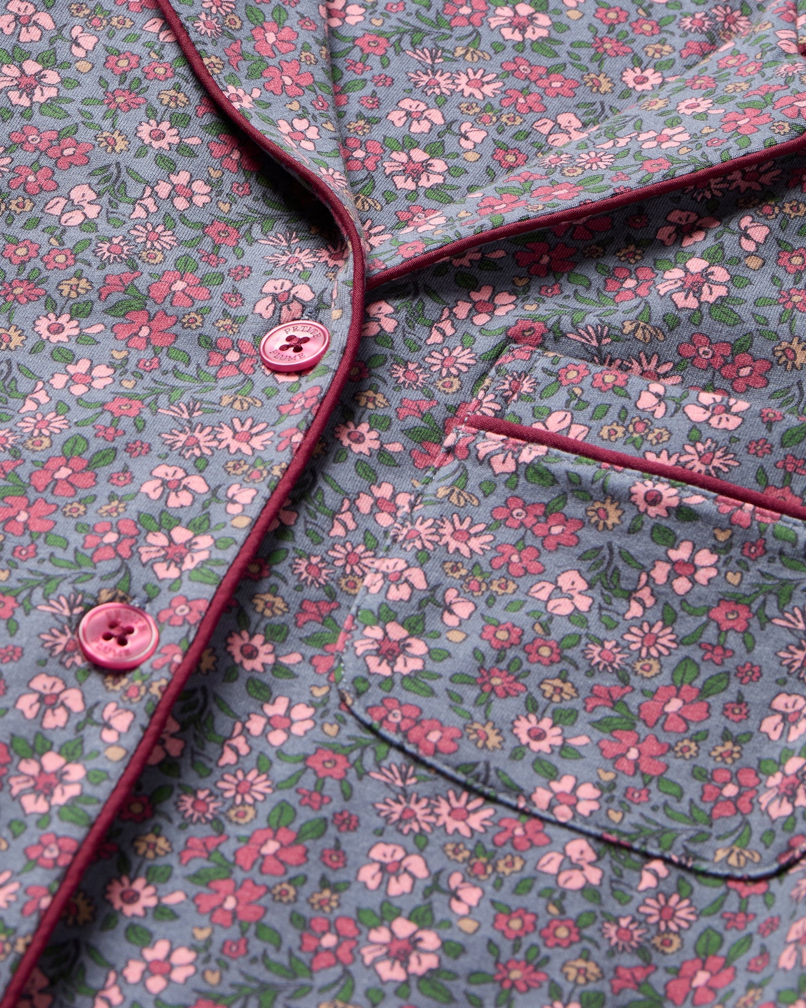 The Women's Pima Pajama Set in Fleurs de Saison features long sleeves and a small floral print in pink, white, and blue on gray. The set includes a button-up top with a chest pocket and matching pants.