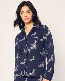 A woman in Petite Plumes Womens Pima Pajama Set in Zebre de Nuit, featuring a navy zebra print and made of Peruvian Pima cotton with buttons and a collar, stands against a neutral backdrop. She sports long, wavy dark hair and has a calm expression.
