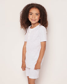 A young child with curly hair smiles against a light background, dressed in Petite Plumes Kids Pima Snug Fit Pajama Short Set in Grey Stars. The white short-sleeved shirt and shorts feature a subtle pattern and neckline buttons, crafted from soft Pima Cotton for comfort.
