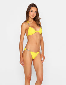 Model wearing the 'Raja Triangle Top - Sunshine' bikini, a vibrant yellow triangle top adorned with large crystal embellishments on the straps. She stands against a white wooden wall, adjusting the matching bottoms and looking down with wavy hair cascading over her shoulder