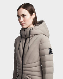 A woman with straight brown hair wears the Rudsak HANNA - 8123562 Cement RDS-certified light beige quilted down jacket with a hood, black drawstrings, and a large R patch on the upper sleeve, against a plain light background.