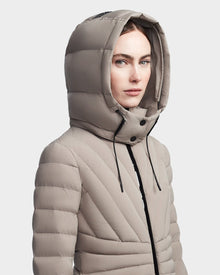 A woman models the Rudsak HANNA - 8123562 Cement, a light taupe hooded down jacket with black drawstrings. Featuring horizontal and diagonal quilting, this winter coat offers stylish warmth against a plain light background.