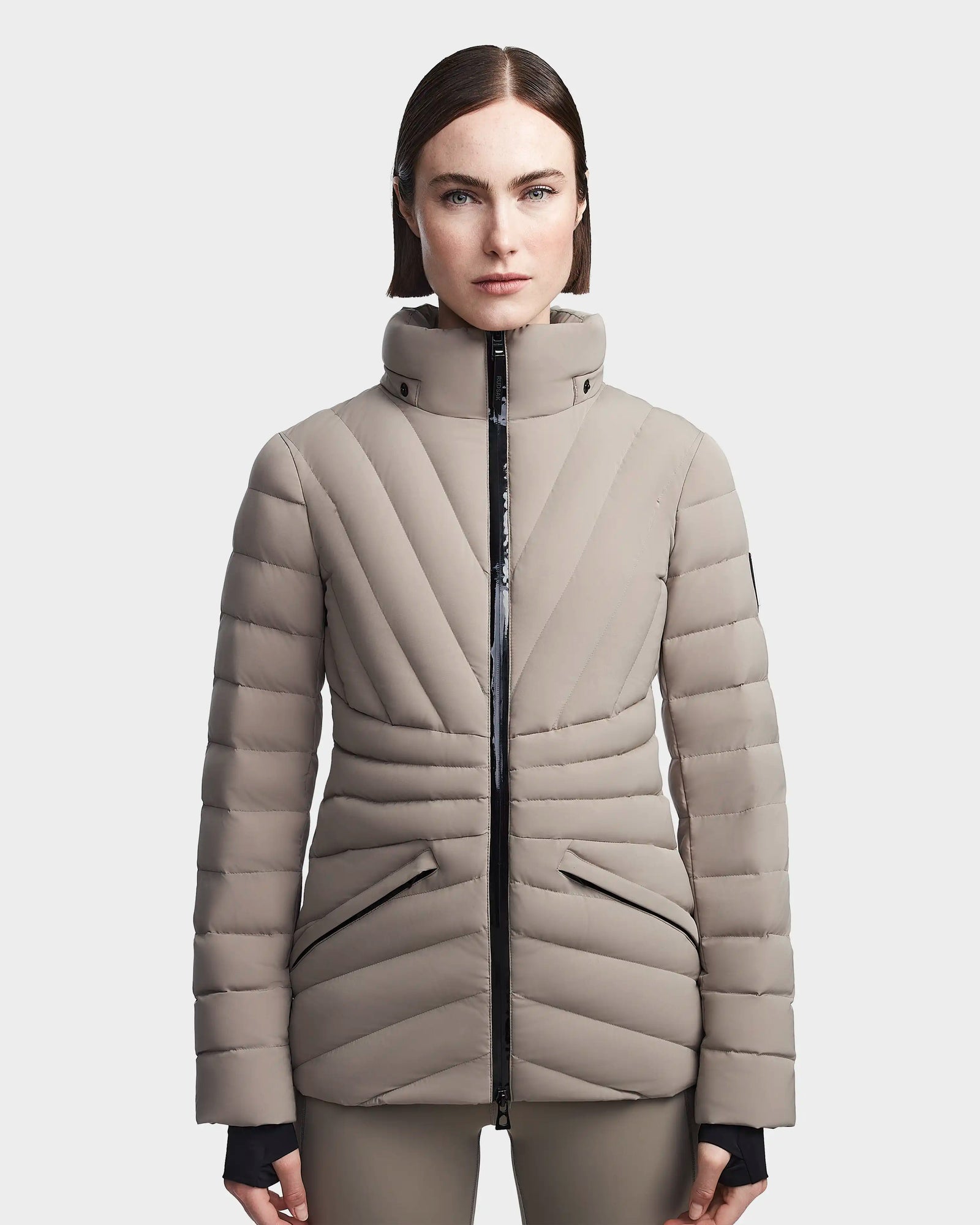 A woman with straight brown hair wears the Rudsak HANNA - 8123562 Cement, a light beige down jacket with a high collar and black zipper, standing forward against a plain white background.
