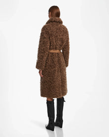 A person stands facing away, wearing the Rudsak KAMILAH - 8124827 Camel womens faux fur trench coat with a vegan leather belt and black knee-high boots against a plain white background.