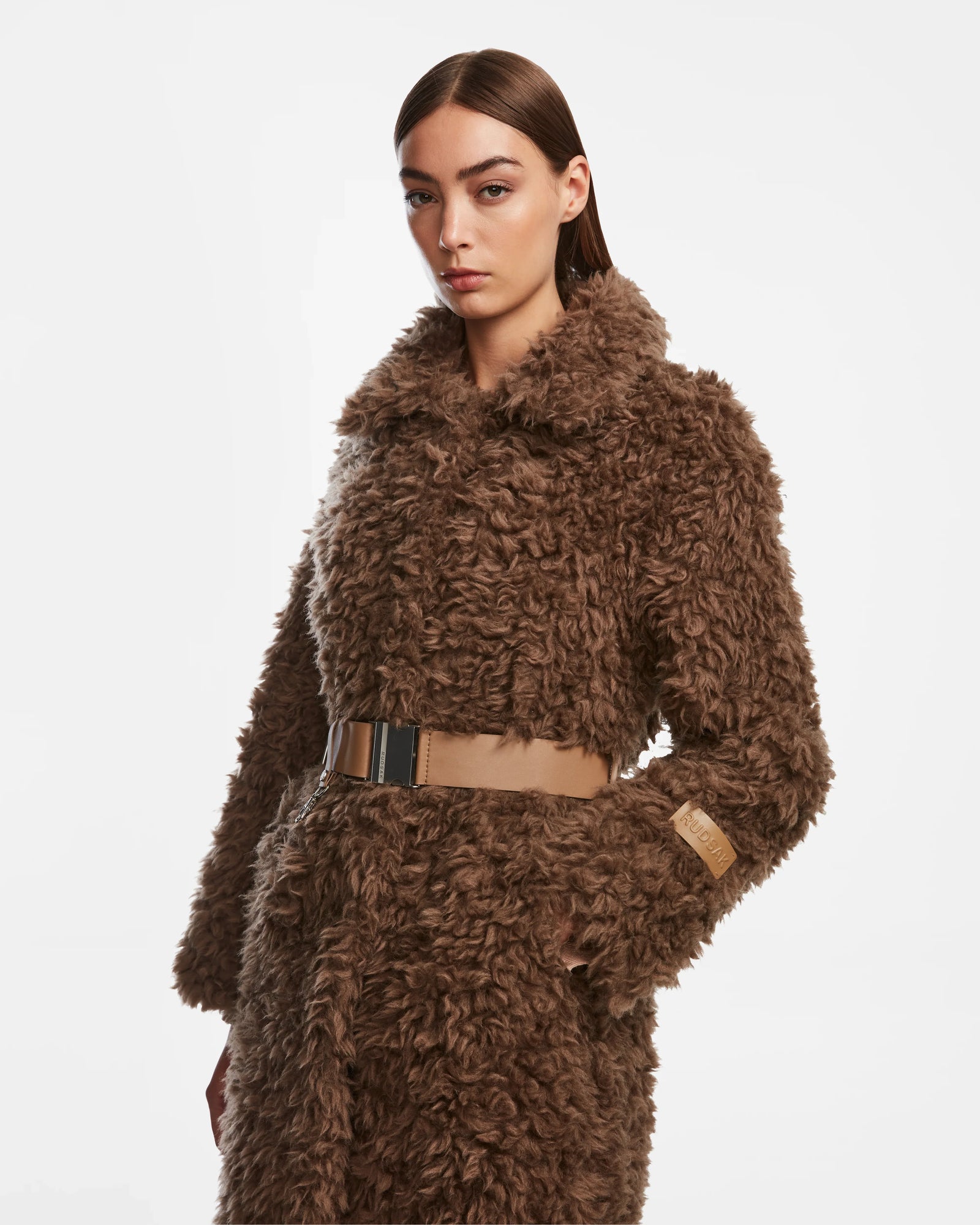 A woman with straight brown hair models the KAMILAH - 8124827 Camel faux fur trench coat by Rudsak, featuring a wide collar and vegan leather belt, standing against a plain white background.