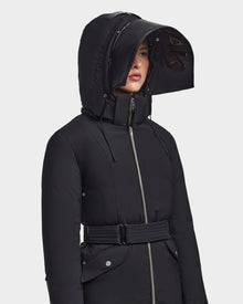 A woman stands against a light background wearing the RUDSAK MILENA - 8122536 BLACK, a long recycled Stormshell winter coat with a structured hood, front zipper, and belted waist.