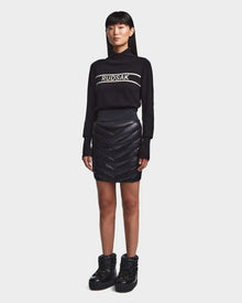 A woman faces forward in a black Rudsak turtleneck sweater, the MARZI - 8523528 Black chevron quilted winter skirt, and black quilted ankle boots, posed against a plain light gray background.
