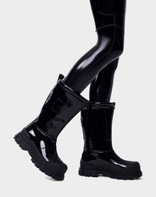 A person wearing Rudsaks KRISTAL HI - 8222510 BLACK high-gloss womens boots with chunky soles and black leggings stands with one foot slightly raised against a light gray background.