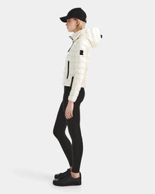 A person in profile wears the Rudsak MELISSA - 8124031 Iridescent Eggshell lightweight down puffer, a black cap, black leggings, and black sneakers against a plain light background.