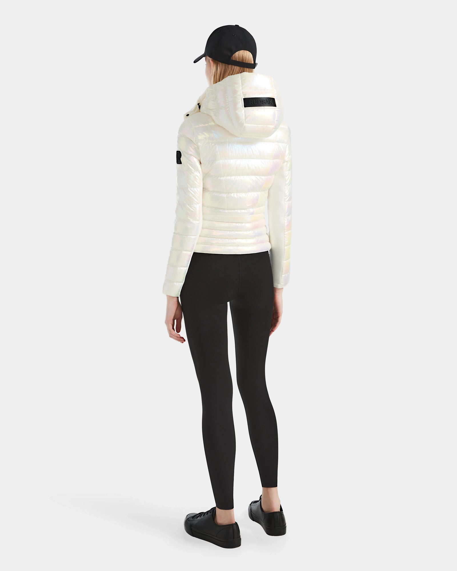 A person stands facing away, wearing the Rudsak MELISSA - 8124031 Iridescent Eggshell women’s lightweight down puffer jacket with a hood, paired with black leggings, sneakers, and a baseball cap against a plain light background.