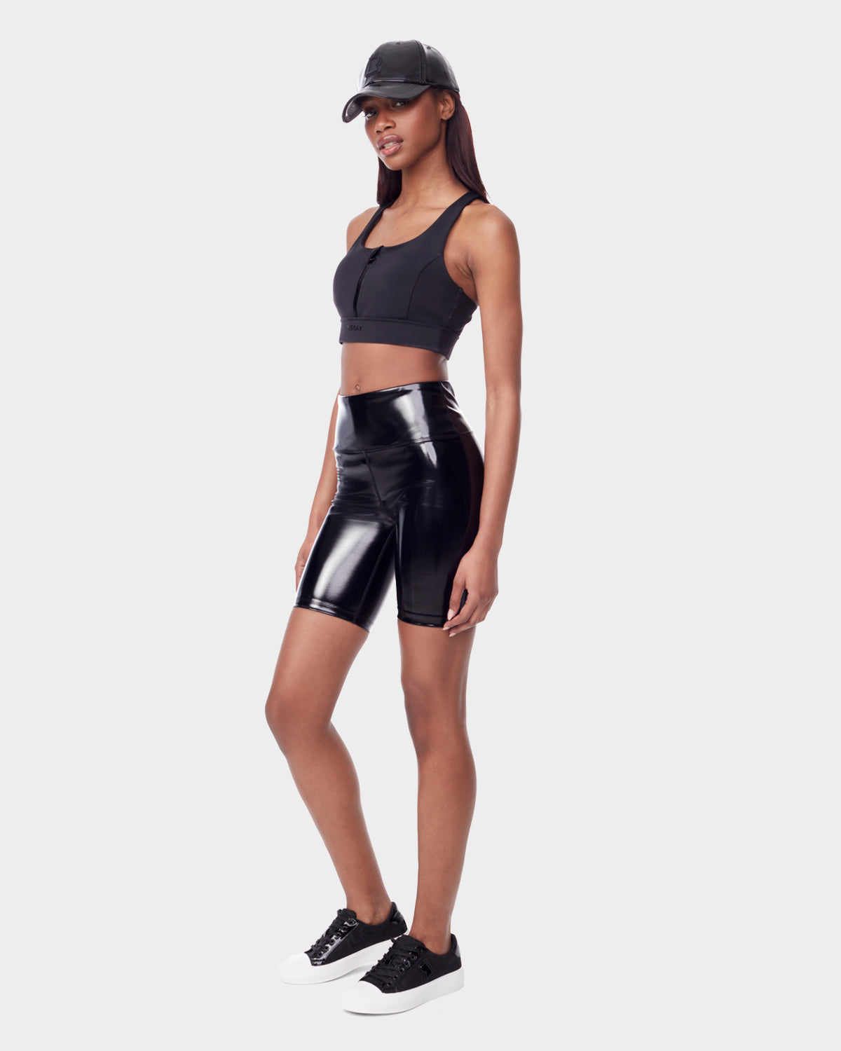 A woman in Rudsak’s BRIA - 8523016 BLACK vegan leather biker shorts, black crop top, sneakers with white soles, and a black cap stands against a light gray background, showcasing sustainable fashion.
