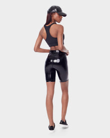 A woman faces away, wearing the BRIA - 8523016 BLACK by Rudsak—a black racerback crop top and high waist vegan leather biker shorts—with black sneakers and a cap. She looks to the side, hand on hip, against a white background.