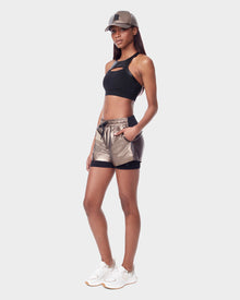 A woman stands confidently against a white background wearing the Rudsak RYLEE - 8523001 METALLIC CHOCOLAT metallic glossy running shorts, a black crop top, white sneakers, and a matching metallic cap, with one hand in her pocket.
