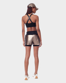 A woman stands facing away, wearing Rudsak’s RYLEE - 8523001 METALLIC CHOCOLAT shiny women’s running shorts with metallic glossy fabric and spandex inner short, paired with a black crisscross sports bra and white sneakers against a light background.