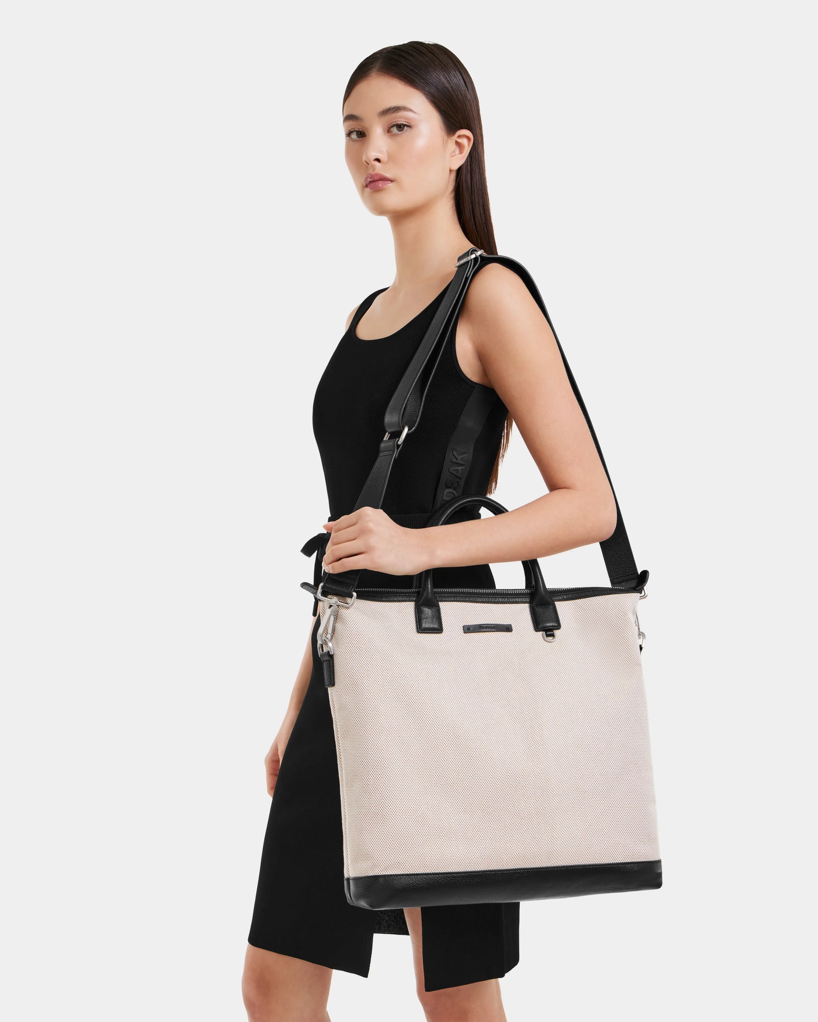 A woman in a sleeveless black dress stands against a white background, carrying the Rudsak DEXTER - 9324513 Vanilla tote, featuring both shoulder and hand straps—a versatile accessory perfect for any occasion.
