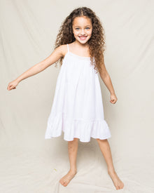 A cheerful young girl with curly hair wears Petite Plumes Girls Twill Lily Nightgown in White, its yarn-dyed cotton adding charm as she smiles barefoot against a light backdrop, her right arm slightly raised.