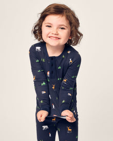A child, smiling in bedtime fun, wears Petite Plumes Kids Pima Snug Fit Pajama Set in Arctic Antics. The dark pajamas feature bears, foxes, and penguins on a light background with trees. Theyre playfully stretching the soft Pima cotton long sleeve top.