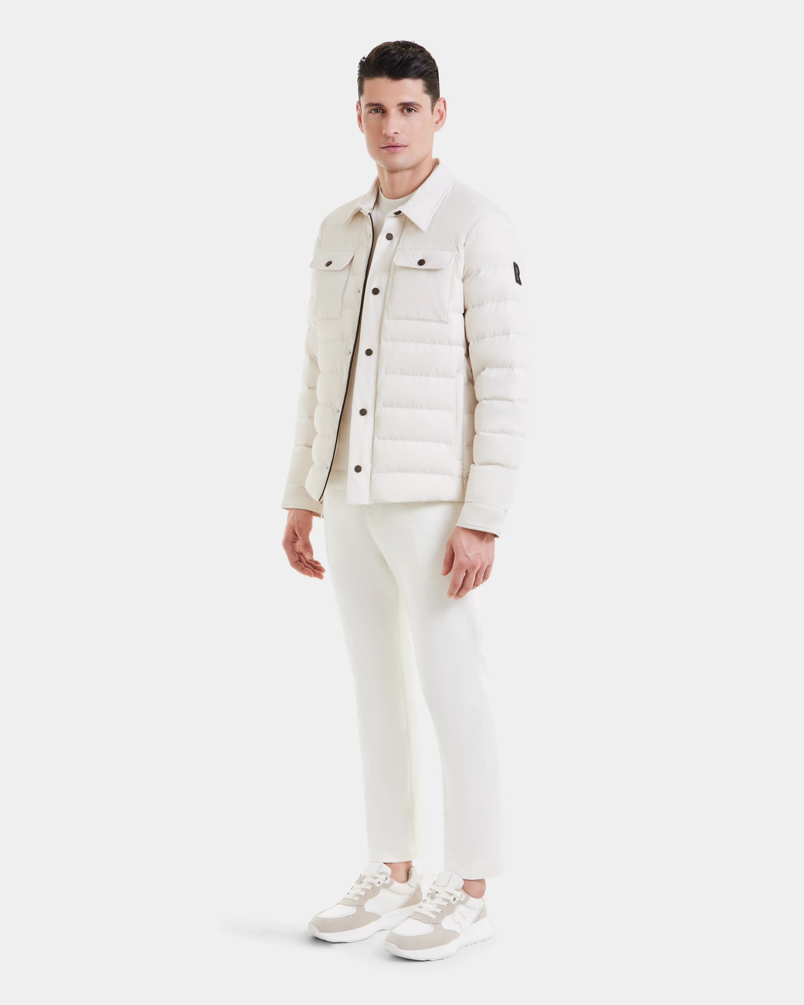 A man stands against a plain white background wearing the Rudsak AZIEL - M124521 Vanilla weather-resistant jacket, paired with white pants and sneakers. He looks forward neutrally, arms relaxed by his sides.