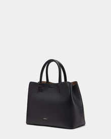 The Rudsak CATIRA S - 8325006 Black is a women’s leather tote bag featuring two handles, minimal stitching, a subtle gold logo near the bottom, and a removable shoulder strap, displayed on a plain white background.