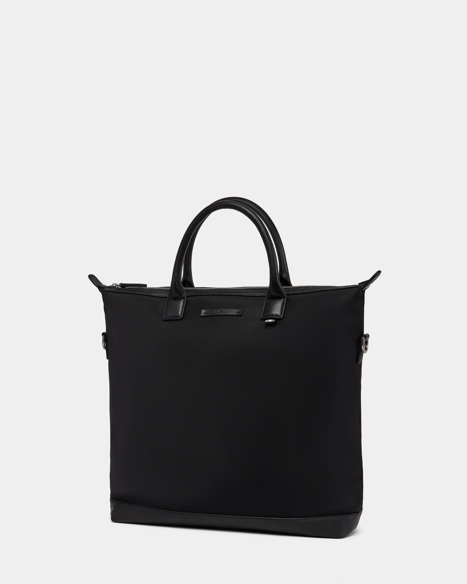 The Rudsak DEXTER - 9324513 Black is a minimalistic black canvas tote bag with two short handles and a zipper closure, showcased against a plain light gray background.