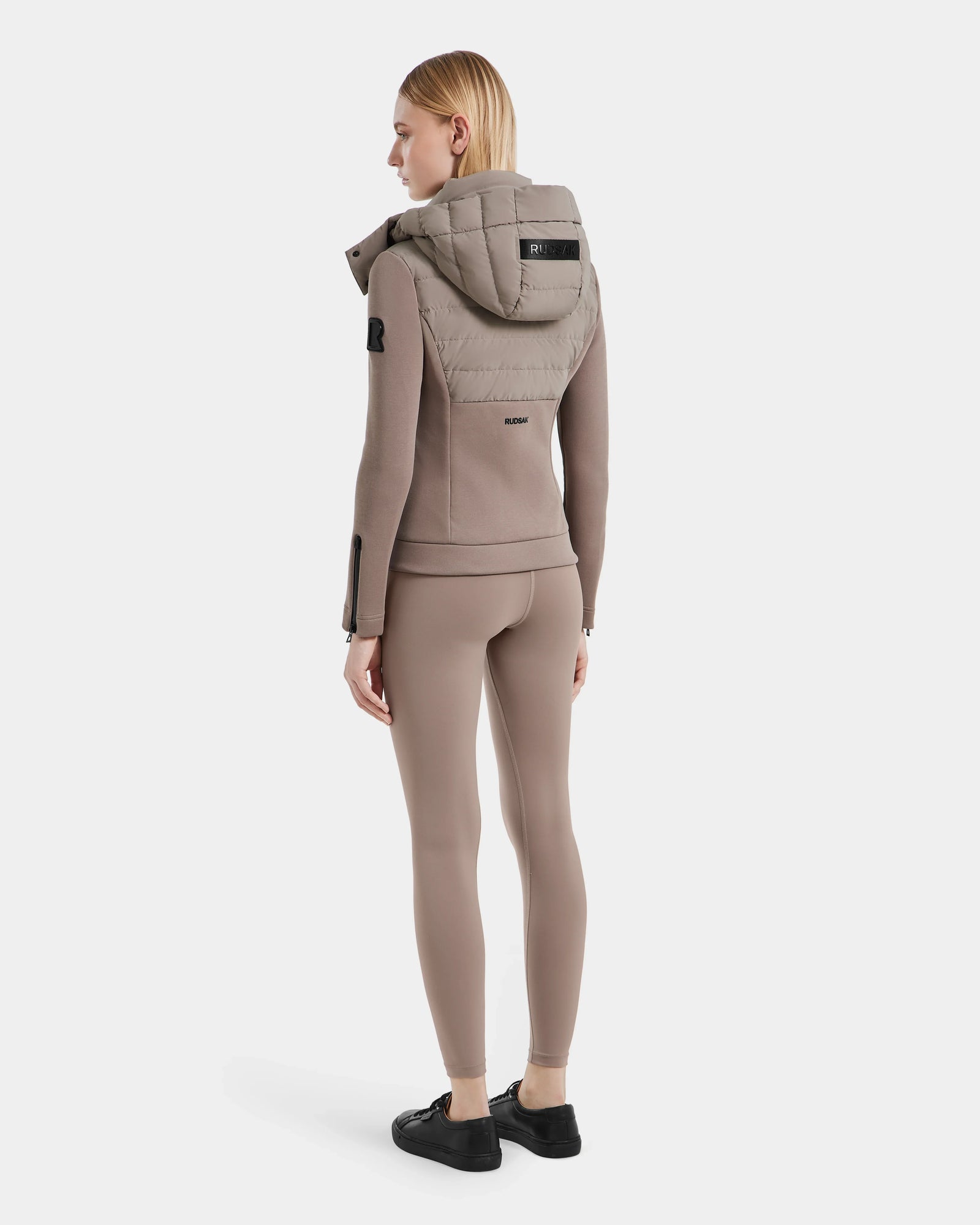 A woman faces away, wearing the Rudsak MABS - 8124825 Cement lightweight taupe short jacket with matching fitted leggings and black sneakers, set against a plain white background.